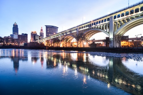 A city skyline at dusk with a bridge reflecting in the water below, showcasing lights and buildings in Cleveland.