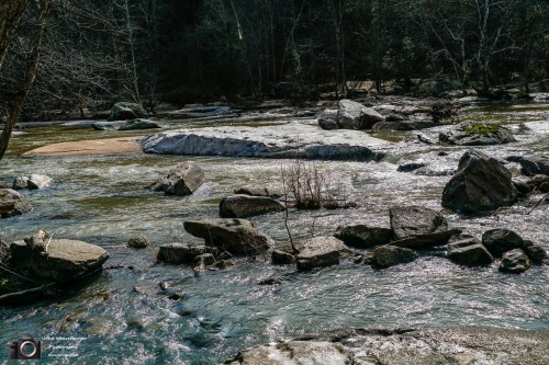 A serene river scene with smooth rocks and gentle water flow, surrounded by trees and a clear blue sky.