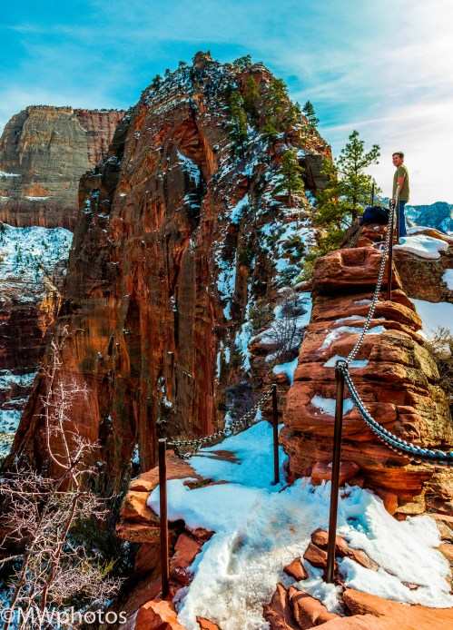 A person stands on a rocky ledge with a chain railing, overlooking a snowy canyon landscape in Zion National Park.