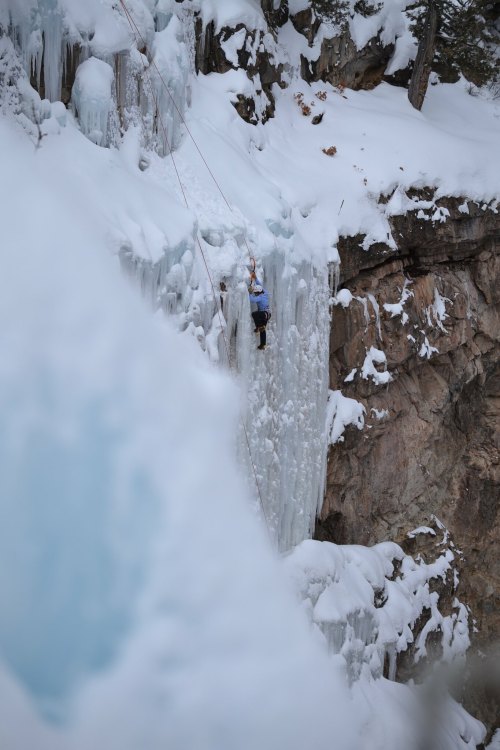 A climber ascends a frozen waterfall surrounded by snow-covered rocks and ice.
