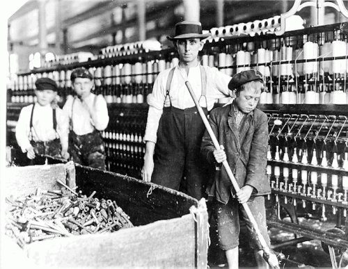 Black and white photo of young boys working in a textile factory, with one boy pushing a cart and others in the background.