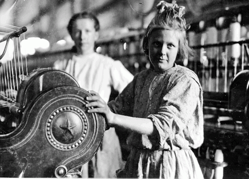 Two young girls working at a textile mill, one operating a loom, in a historical black-and-white photograph.