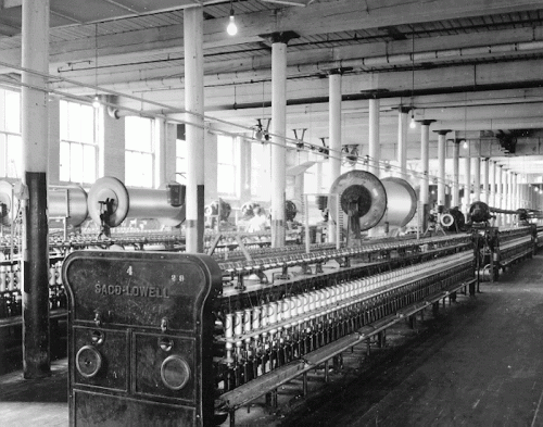 Black and white image of a textile mill interior with rows of spinning machines and large windows.