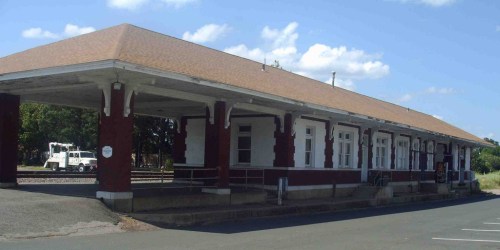 A historic train station with a large overhang, featuring red brick and white trim, set against a blue sky.