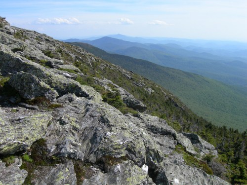 A rocky mountain slope with green hills and distant blue mountains under a clear sky.