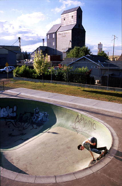 A skateboarder performs a trick in a graffiti-covered bowl, with a grain elevator and trees in the background.