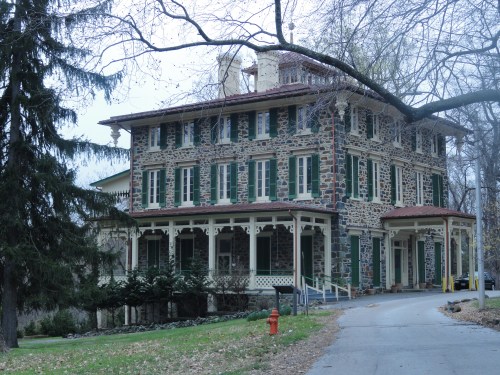 A historic stone house with green shutters, surrounded by trees and a winding driveway, on a cloudy day.