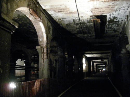 Dimly lit underground corridor with arched ceilings and weathered walls, creating an eerie atmosphere.
