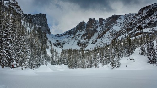 A snowy landscape with towering mountains and evergreen trees under a cloudy sky. A person is visible in the distance.