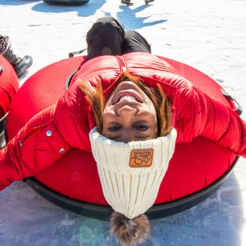 A person in a red jacket and white beanie smiles while lying on a large, round inflatable tube on snow.