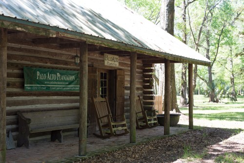 A rustic wooden cabin with a porch, rocking chairs, and a sign for Palo Alto Plantation, surrounded by greenery.