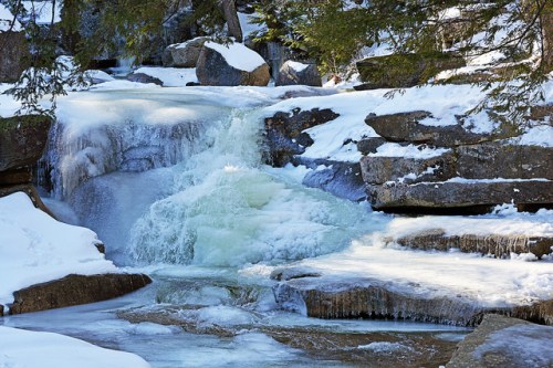 A frozen waterfall cascades over rocks, surrounded by snow-covered ground and evergreen trees.