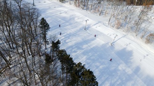Aerial view of a snowy landscape with skiers on a slope surrounded by trees and ski lift poles.