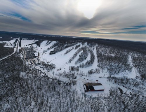Aerial view of a snowy landscape with hills, trees, and a building, under a cloudy sky.