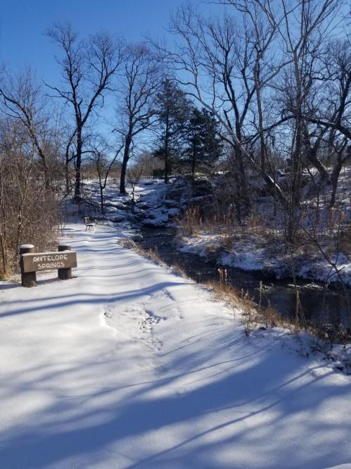 A snowy path beside a stream, with bare trees and a sign for "Art Lodge Springs" in the foreground.