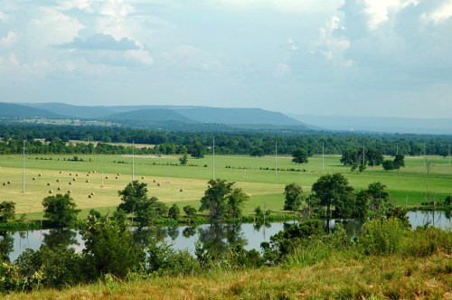Lush green fields with hay bales, a calm pond, and distant mountains under a cloudy sky.