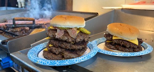 Two stacked burgers on paper plates, one with multiple patties, cheese, bacon, and pickles, with a grill in the background.