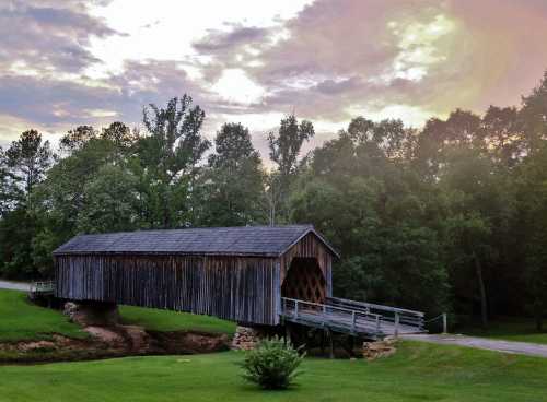 A rustic covered bridge spans a small creek, surrounded by lush greenery and a cloudy sunset sky.