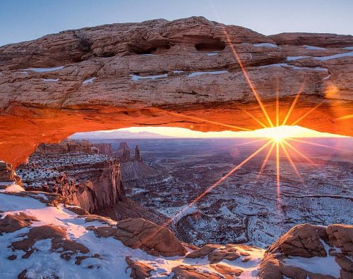 Sunrise shining through a natural rock arch, illuminating a snowy landscape and distant rock formations.