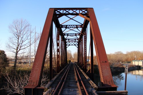 Rusty metal train bridge spanning a calm river, with trees and power lines in the background under a clear blue sky.