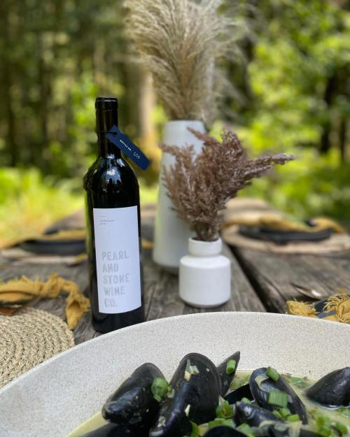 A bottle of Pearl and Stone Wine Co. wine beside a dish of mussels, with decorative vases and greenery in the background.