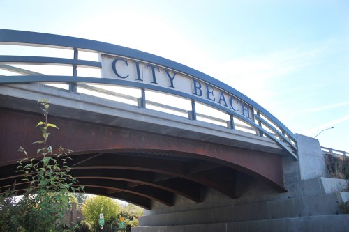 A bridge with the sign "CITY BEACH" prominently displayed against a clear blue sky. Green foliage in the foreground.