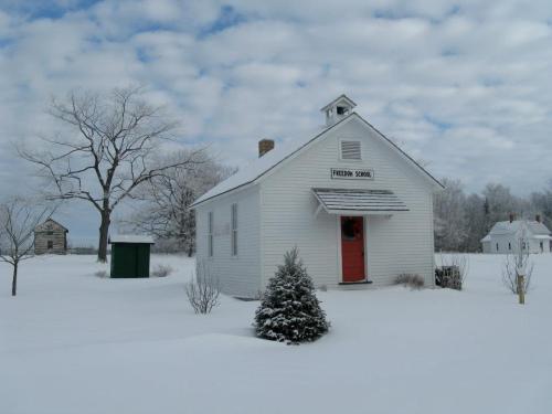 A small white schoolhouse with a red door surrounded by snow, under a cloudy sky, with trees in the background.