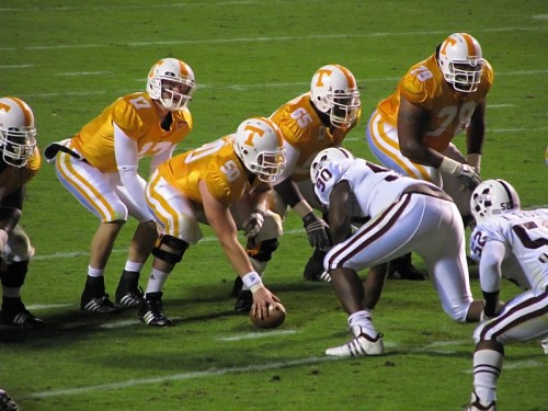 A football team in orange jerseys prepares for a snap, facing an opposing player in white.