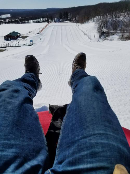 A person sitting on a sled, looking down a snowy slope with trees and a building in the background.
