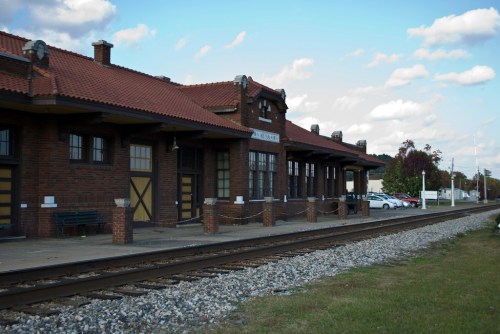 Historic train station with a red-tiled roof, brick facade, and a gravel platform beside railway tracks.