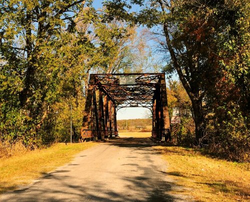 Rusty metal bridge surrounded by trees, leading to an open field under a clear blue sky.