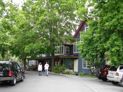 Two people walk along a tree-lined path near a blue house, with parked cars visible in the foreground.