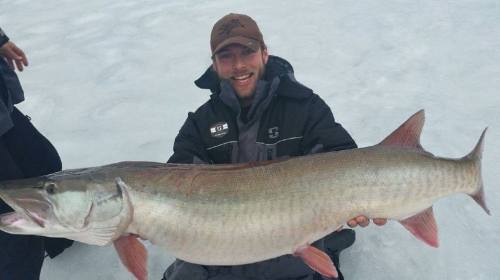 A smiling man holds a large fish while sitting on ice, dressed in winter gear.