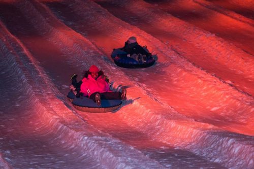 Two people sledding down a snowy hill at night, illuminated by warm orange lights.