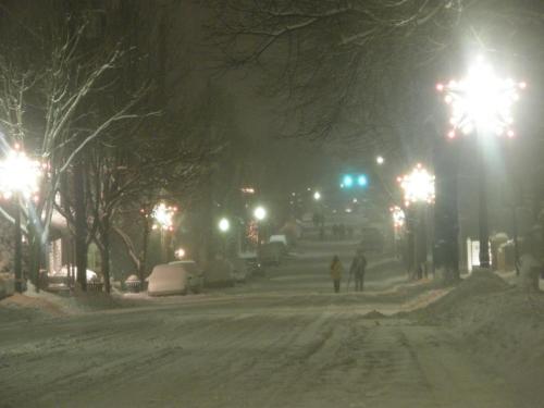 A snowy street at night, illuminated by festive lights, with two people walking in the distance.