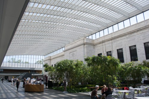 Bright indoor space with a glass roof, featuring greenery, people walking, and a café area.