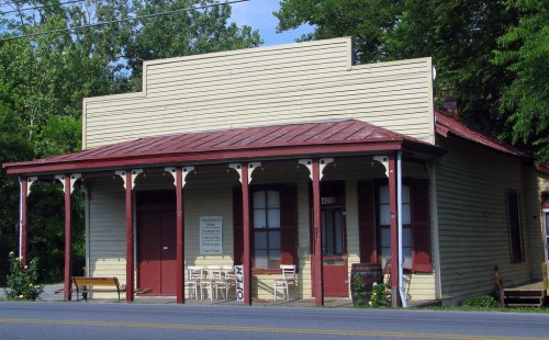 A vintage yellow building with a red roof and porch, featuring white chairs outside and surrounded by greenery.