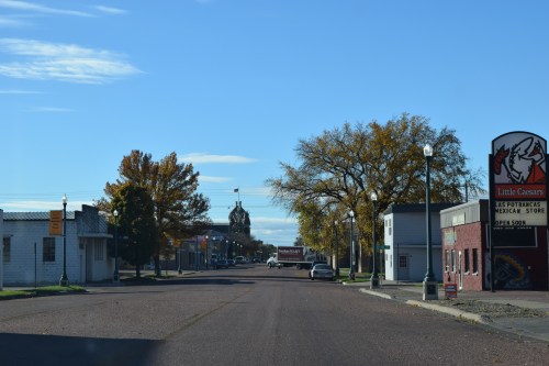 A quiet street scene with storefronts, trees, and a distant clock tower under a clear blue sky.