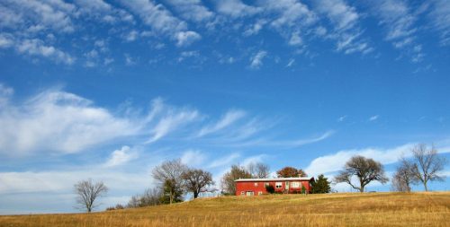 A red house on a grassy hill under a blue sky with wispy clouds and a few bare trees.