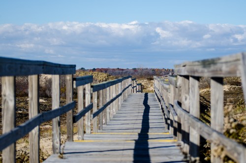 A wooden boardwalk leads through a sandy area, with clouds and trees in the background under a clear blue sky.