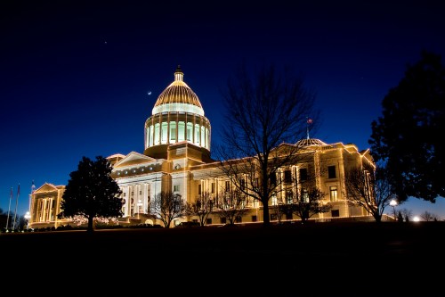 The illuminated dome of a grand building at night, surrounded by trees against a deep blue sky.