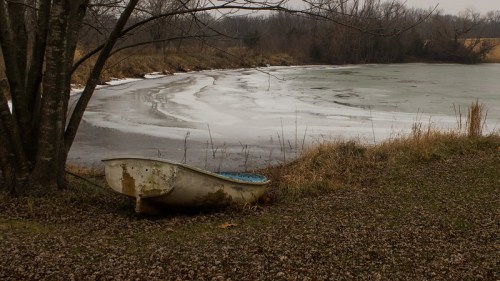 An old, rusty bathtub sits by a frozen pond, surrounded by bare trees and winter grass.