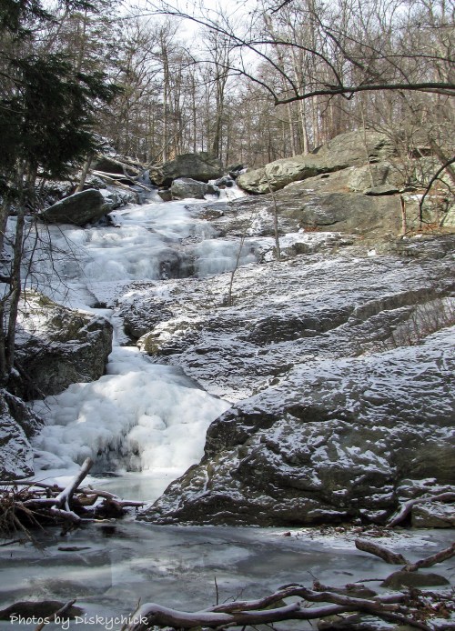 A frozen waterfall cascades over rocky terrain, surrounded by snow-covered trees and a serene winter landscape.
