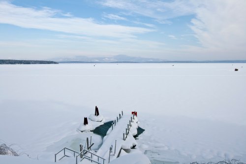 A snowy landscape with a frozen lake and a wooden dock extending into the ice, under a cloudy blue sky.