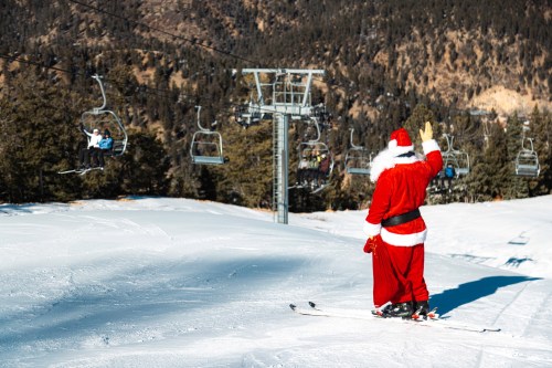Santa Claus in a red suit waves while skiing on a snowy slope, with ski lifts and trees in the background.