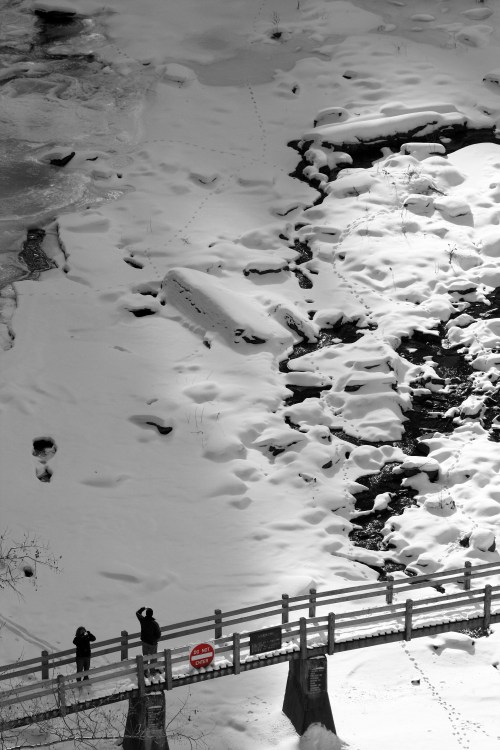 Two people stand on a snow-covered bridge, overlooking a frozen river with footprints in the snow nearby.