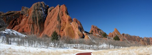 Red rock formations rise against a clear blue sky, with a snowy landscape and bare trees in the foreground.