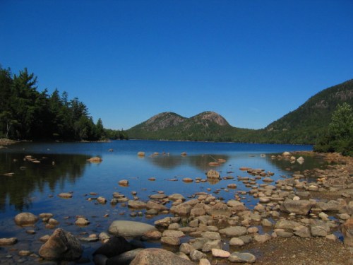 A serene lake surrounded by mountains, with clear blue skies and rocky shores reflecting in the calm water.