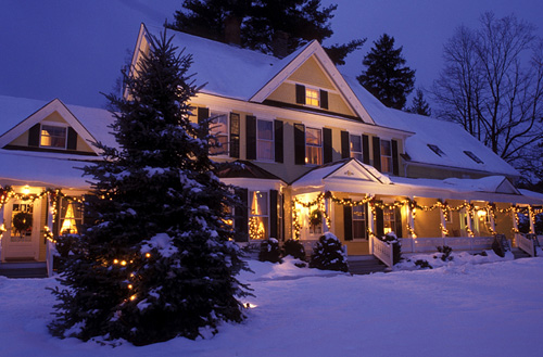 A cozy house decorated with lights, surrounded by snow and a Christmas tree, under a twilight sky.