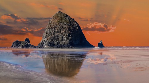 A rocky formation stands on a beach at sunset, reflecting in the wet sand with colorful clouds in the sky.